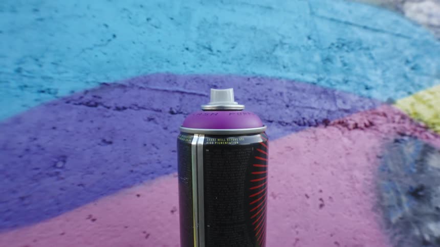 A can of spray paint with a purple cap and a black and orange design standing against a painted wall
