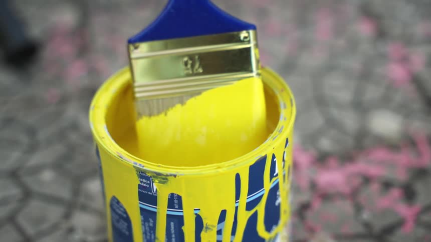 A paintbrush with blue bristles is dipping into a can of yellow paint