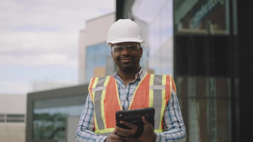 A worker in a white hard hat and high visibility safety vest holding a tablet standing in front of a building