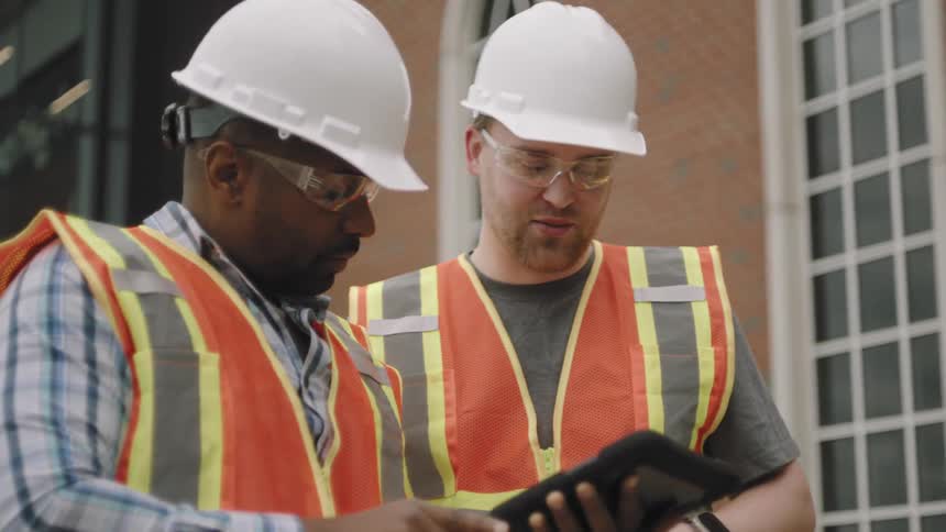 Two workers with protective helmets converting between them and holding a tablet