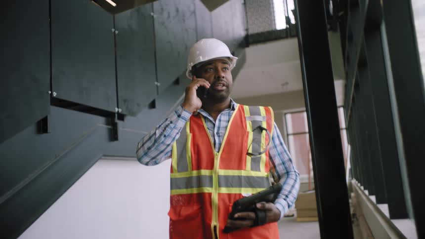 Man holding a tablet and talking on his cell phone while wearing a protective vest and helmet
