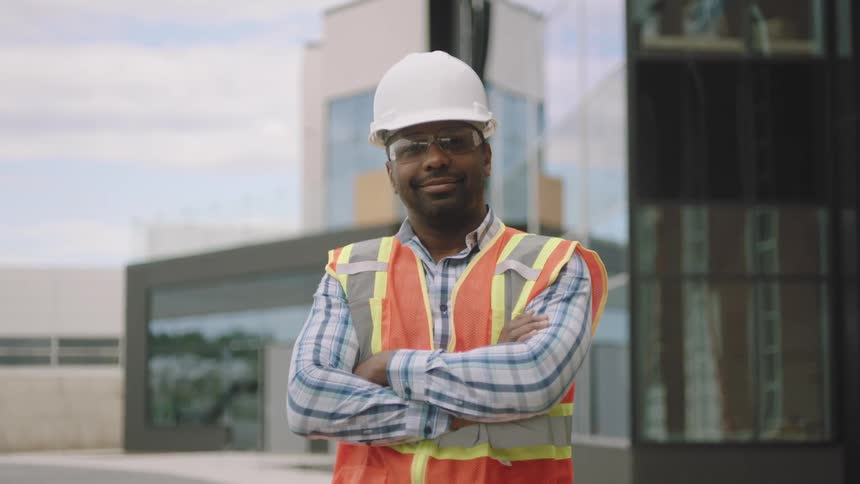 A confident construction worker wearing a hard hat safety glasses and a reflective vest is standing with arms crossed in front of a modern building