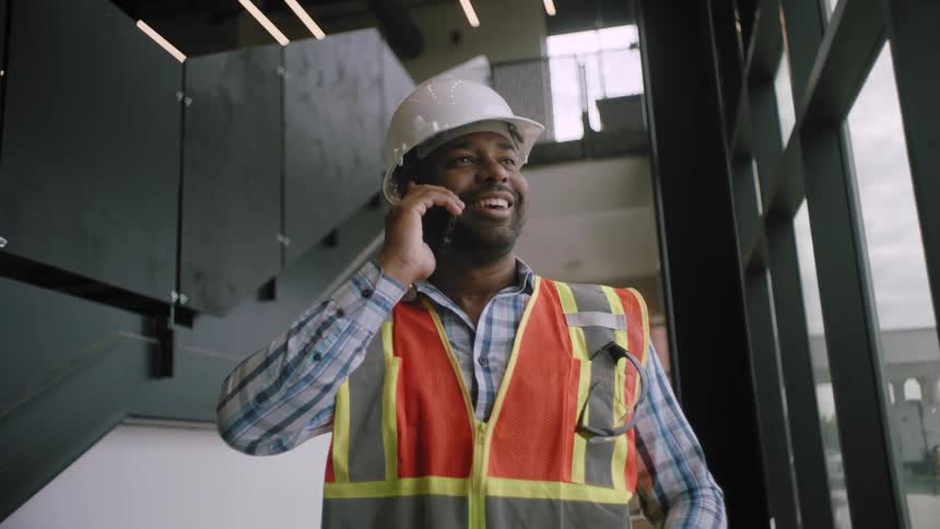 A construction worker wearing a hard hat and a reflective vest is talking on the mobile phone in a building