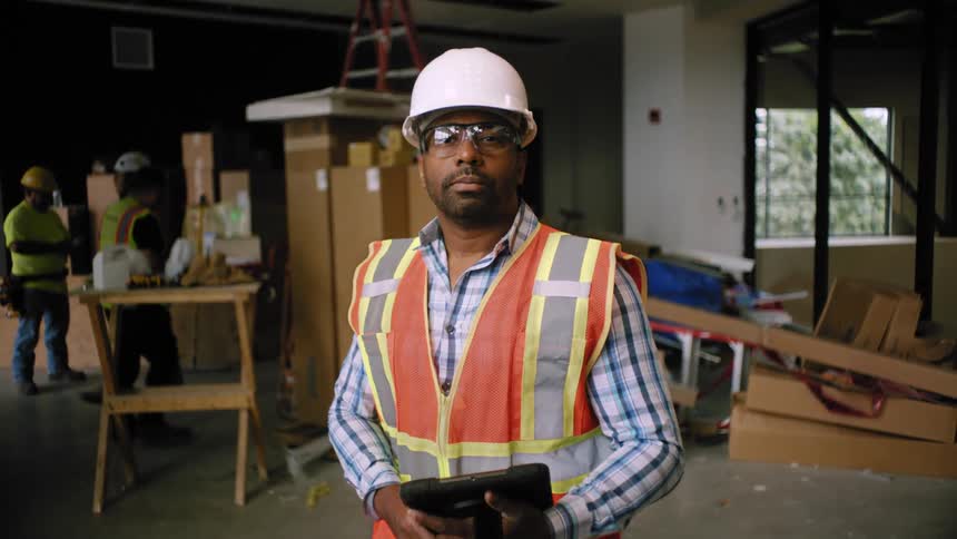 Man in helmet and vest at a construction site indoors holding a tablet