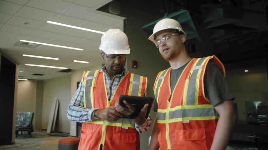 Two men wearing safety helmets and high visibility vests one holding a tablet and discussing work related matters