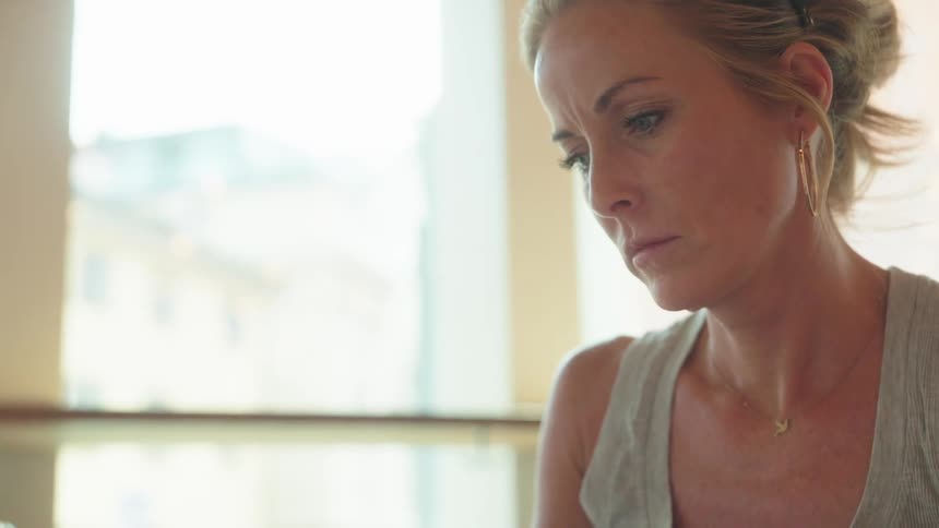 Middle aged blonde woman with hoop earrings and a gray top looks down at laptop screen reading