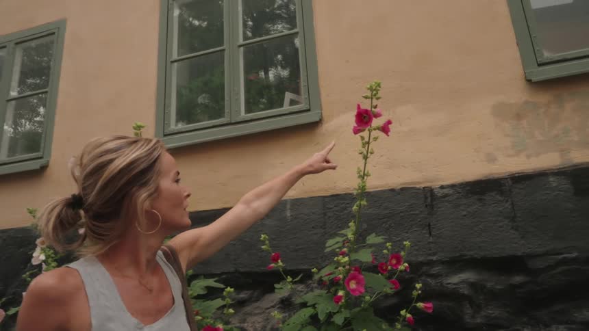 Woman walking and pointing to a pink flower in a flowerbed of a building