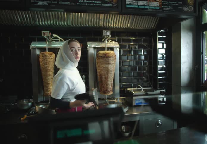 Young woman in white sweatshirt and cap working in a restaurant with meats in the background