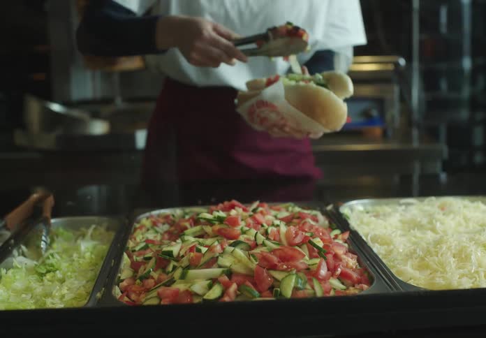 Person adding diced tomatoes to a sandwich using tongs with trays of ingredients in front