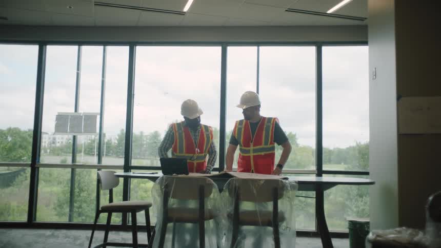 Two men wearing high visibility vests and helmets are standing next to a table talking