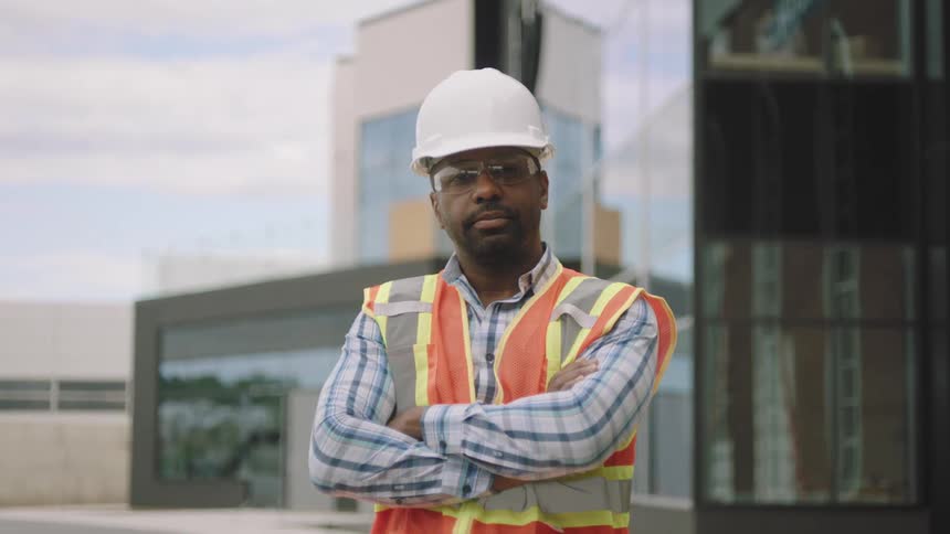 A person in a highvisibility vest and hard hat stands with arms crossed in front of a building