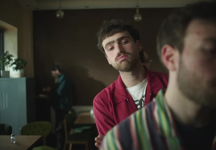 Three men waiting in line in a cafeteria peeking ahead while another man eats in the back of the room