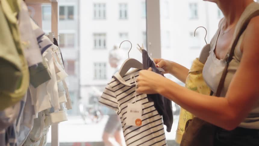 Blonde woman with sunglasses hoop earrings and a gray top looks for baby clothes in a clothing store