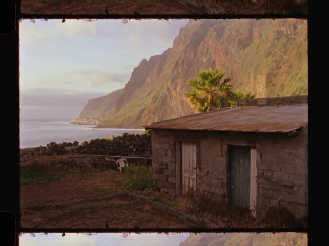 Rustic home with corrugated metal roof in front of towering cliffs and the ocean under a clear sky
