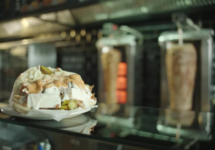 A freshly made dner kebab sandwich filled with sliced meat pickles and drizzled with sauces being placed on a counter in front of rotating dner meat grills