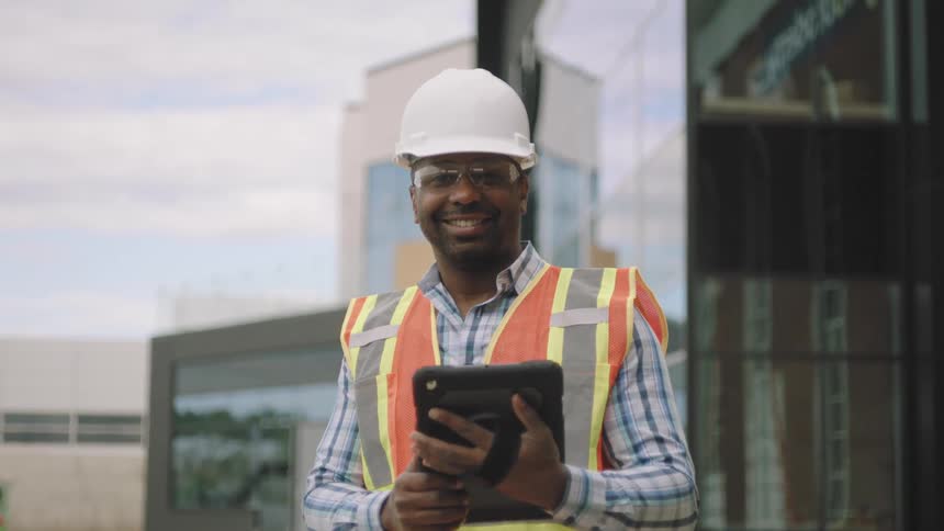 A person in a white hard hat and high visibility vest is holding a tablet
