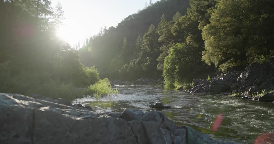 River flowing through a rocky landscape with trees on either side sun shining brightly from behind the hills
