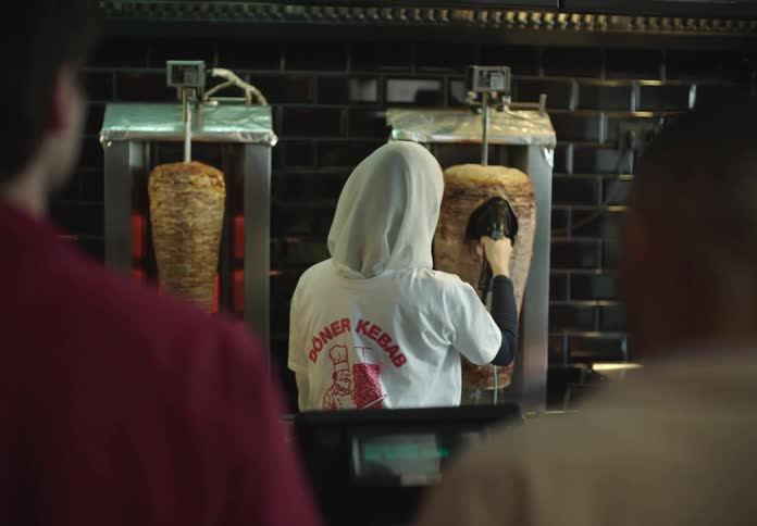Person in a white hoodie standing in front of a vertical kebab rotisserie with large skewers of meat