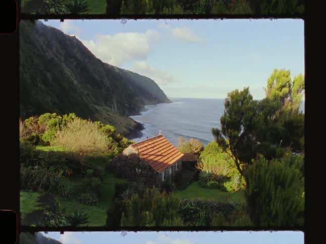 A rustic stone house with a redtiled roof is nestled in the mountains and the ocean in the distance
