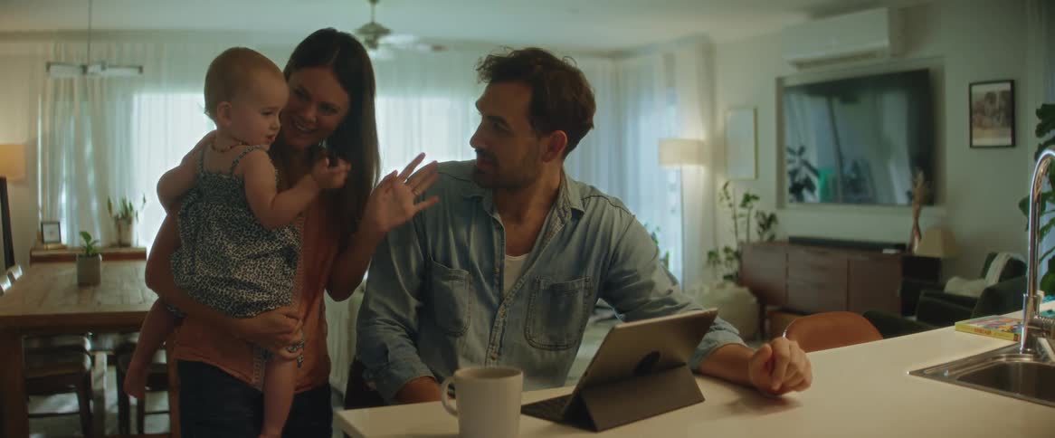 A mother holding her baby standing next to the father as they wave at a tablet screen