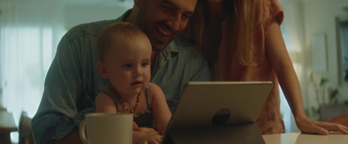 A baby girl on her fathers lap as she smiles and waves her hands at the tablet screen with the mother standing next to them