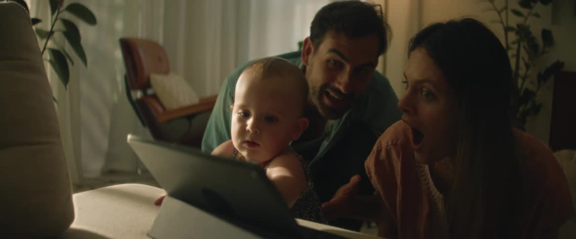A mother and father sit in front of a tablet and try to teach their toddler how to use it