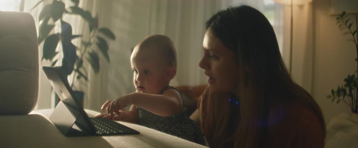 A mother sitting with her baby daughter who is playing with a laptop on the sofa