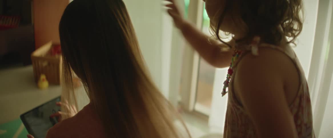 A woman is kneeling down looking at a tablet computer as a little girl standing behind her runs her fingers through her long brown hair