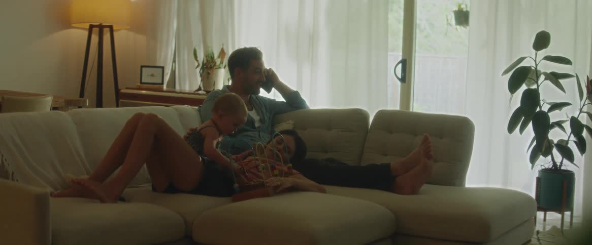 A father and a mother sitting and lying on the living room couch with the father talking on the phone and the mother playing with the baby