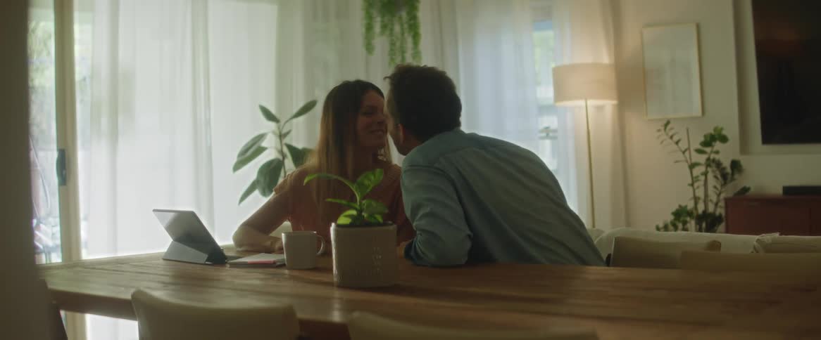 A man and woman sitting at a kitchen table talking smiling then kissing each other