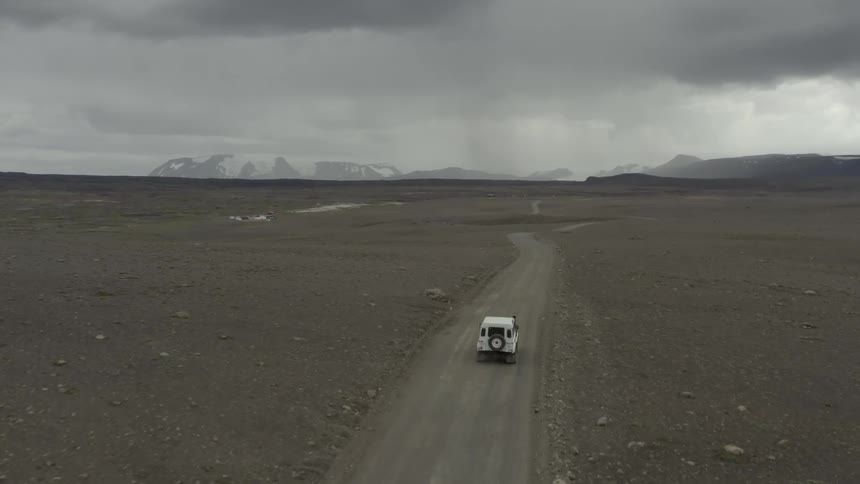 Rain falls over distant mountains as a vehicle drives on a dirt road across a vast plain
