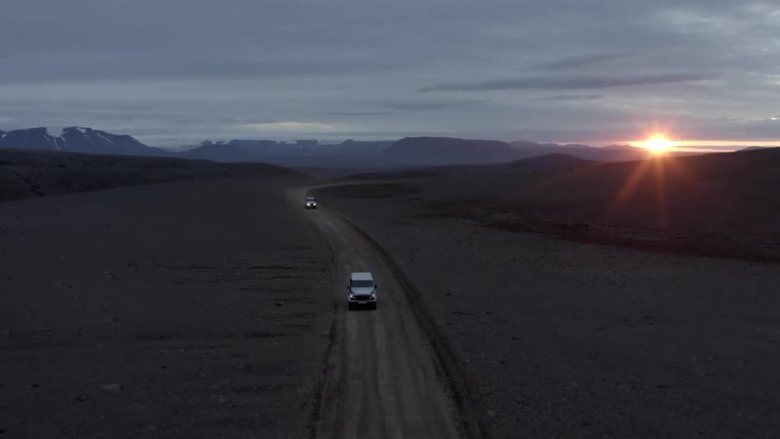 Aerial view of two vehicles traveling on a dirt road with mountains surrounding them and the golden sun on the horizon