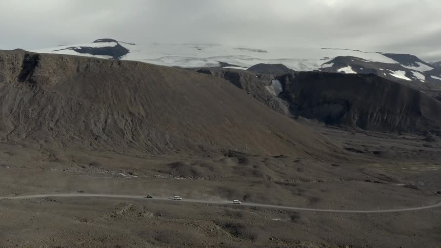 Three vehicles are traveling along a remote road in a mountainous area on a cloudy day