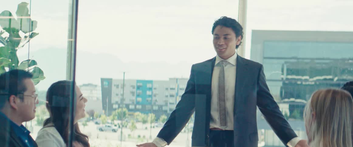 Man in a suit in an office room talking to other colleagues who are sitting