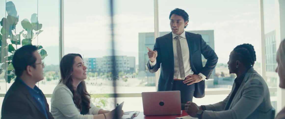 Man standing in a meeting room talking to other people who are sitting around a table