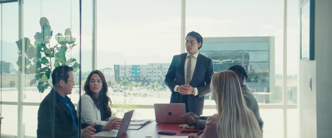 A meeting with four people sitting down and working on laptops and a man standing up who is agreeing with one of the men who is sitting down and talking