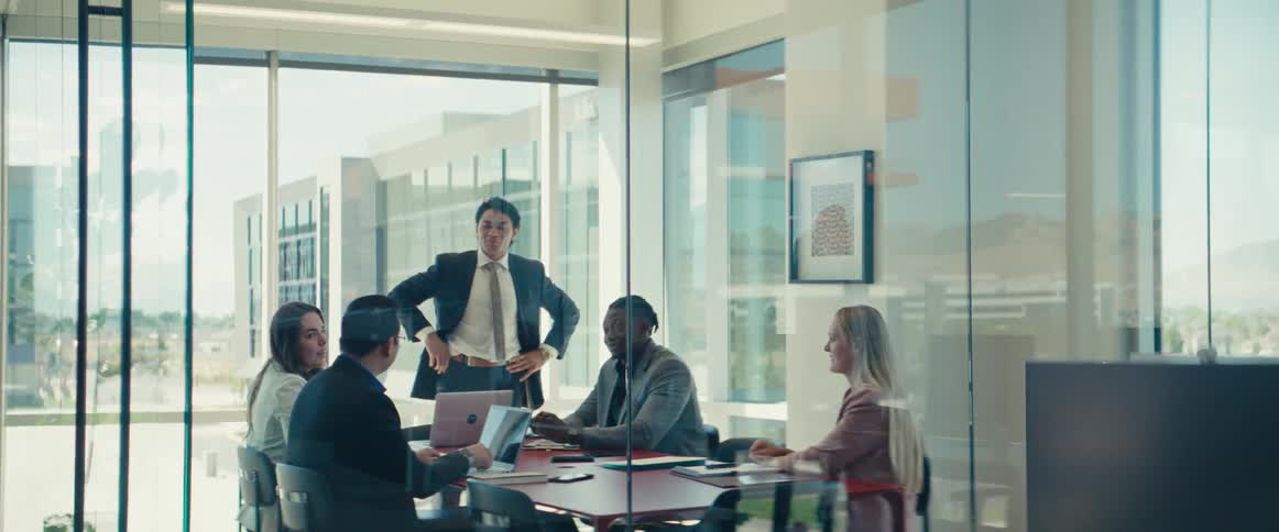 Man in a suit standing talking to a group of people who are sitting at a table in an office meeting