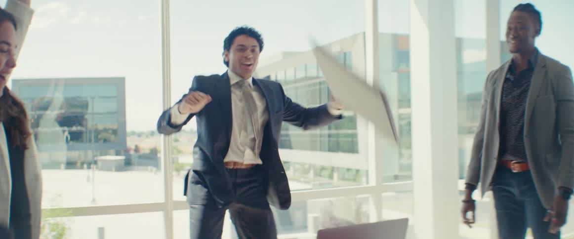 A group of workers in an office celebrate by dancing and throwing papers around