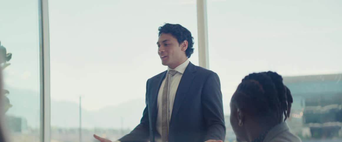 A man in formal attire gives a presentation on a business meeting in a modern office with large windows