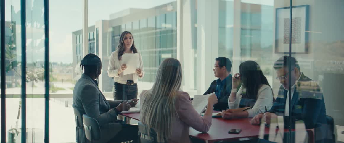 A business meeting in a glass office with one woman standing making a presentation and five people sitting while listening to her and taking notes