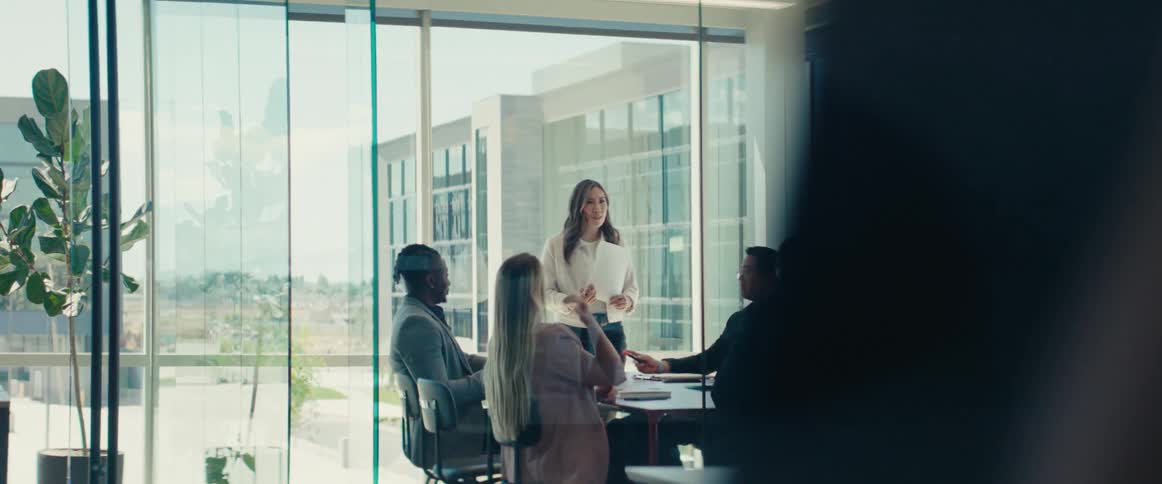 A business meeting in a glass office with one woman standing and five people sitting