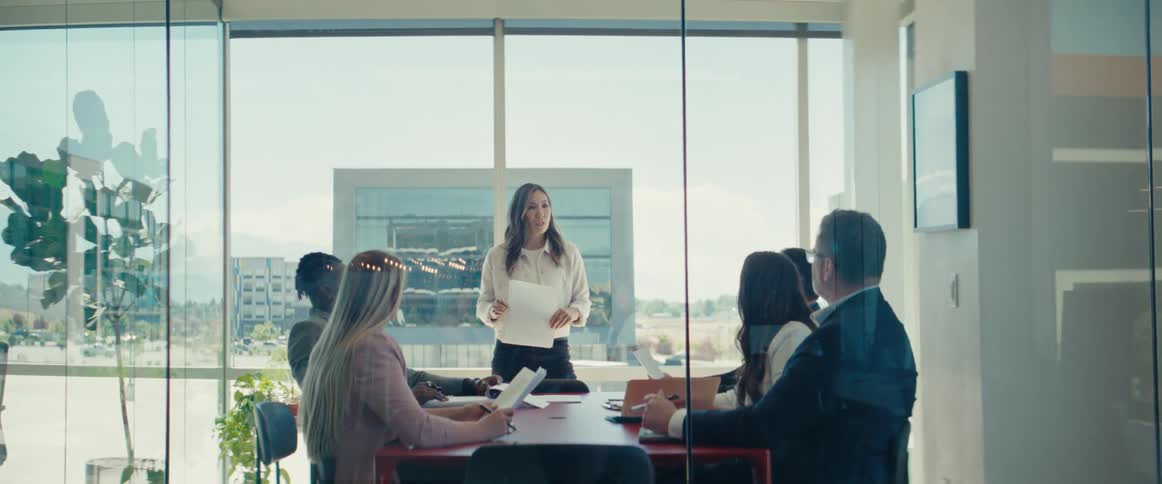 A business meeting in a glass office with one woman standing up to give a presentation while the others watch
