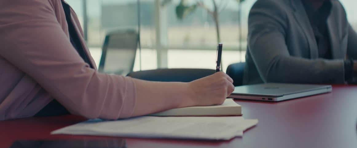 A woman with blonde hair wearing a pink top is seated at a table writing on a notebook with a pen