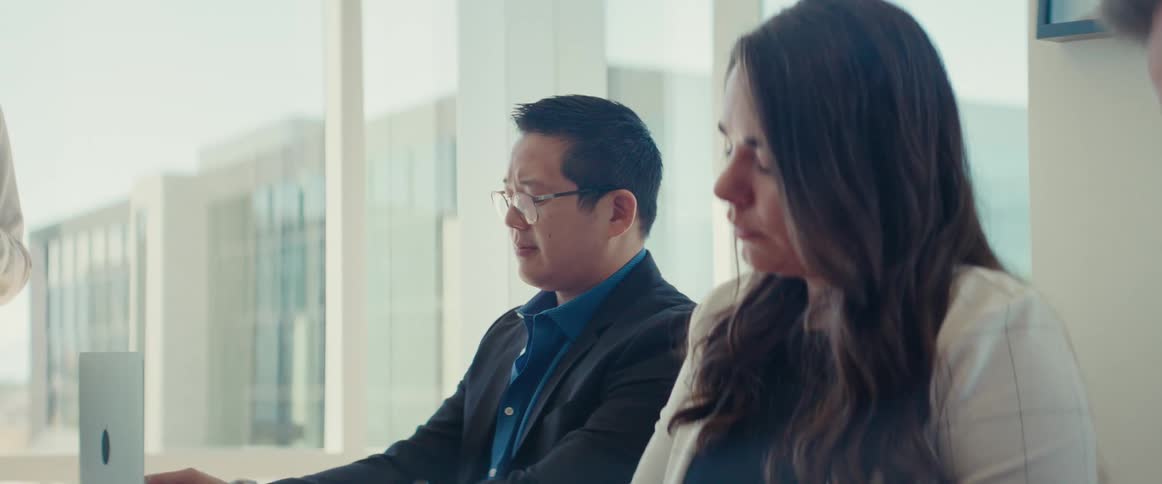 A group of people are seated around a table in an office listening to someone present
