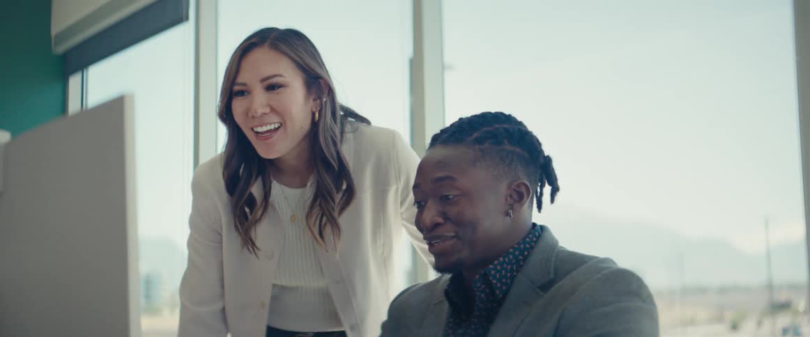 Woman in an office environment talking to a man who is sitting in front of a computer