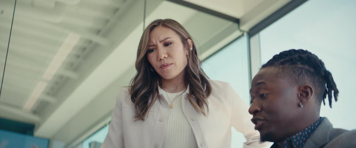 An asian woman walks up to a black office employee working on a laptop and listens to him explain something