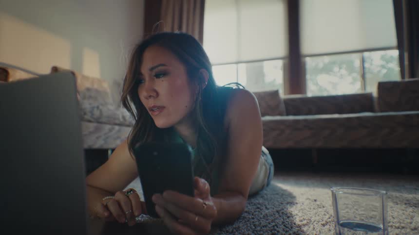 An asian woman lies on the floor while holding her smartphone and working on her laptop