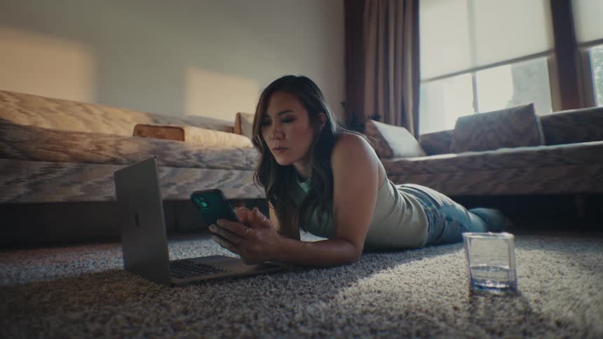Woman lying on living room carpet typing on laptop and interacting with smartphone
