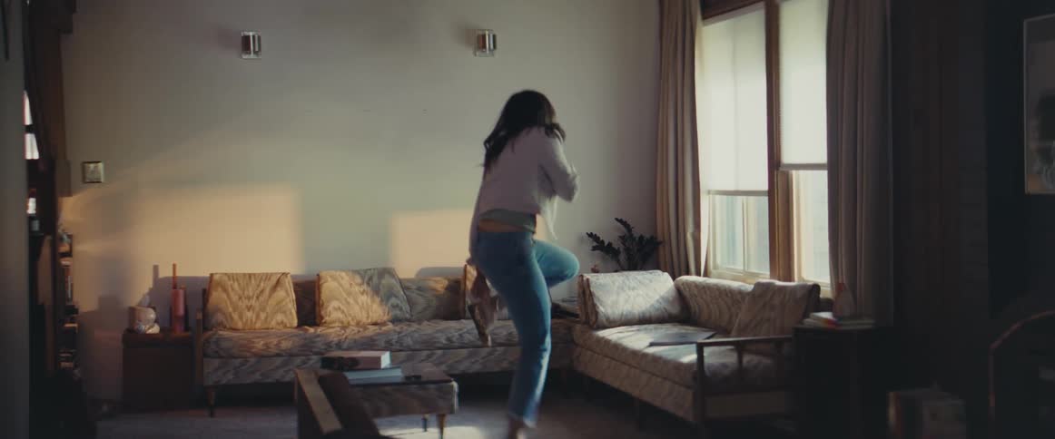 A young woman puts a shoe on while hopping around and then quickly cleans a living room