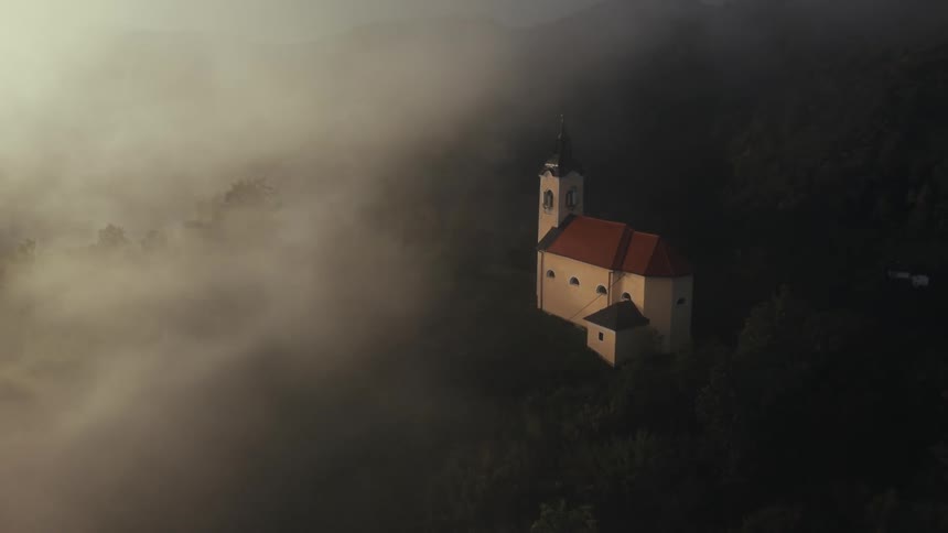 Aerial view of a church surrounded by trees partially shrouded in mist set against rolling hills